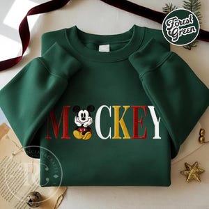 以下が含まれることがあります： 赤、黄、白で「ミッキー」と刺繍されたフォレストグリーンのスウェットシャツ。「M」の文字にはミッキーマウスの刺繍画像が施されています。スウェットシャツは長袖です。