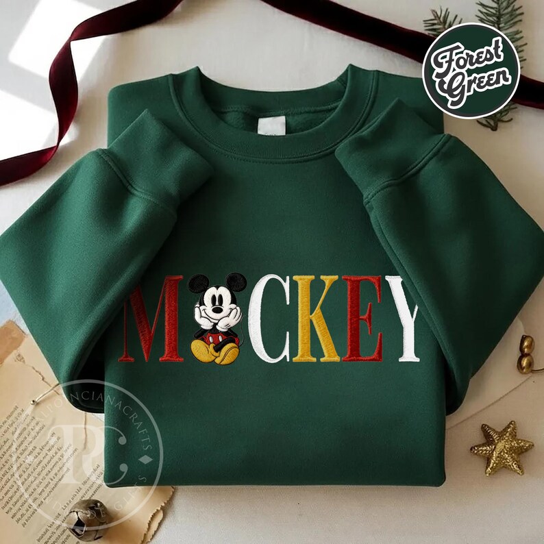 以下が含まれることがあります： 赤、黄、白で「ミッキー」と刺繍されたフォレストグリーンのスウェットシャツ。「M」の文字にはミッキーマウスの刺繍画像が施されています。スウェットシャツは長袖です。