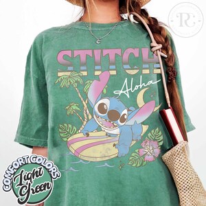 Puede incluir: Camiseta verde claro con un gr&aacute;fico de Stitch surfeando, la palabra "STITCH" en rosa y gris, y la palabra "Aloha" en blanco. La camiseta tambi&eacute;n presenta una palmera, una luna y una flor. Las palabras "Comfort Colors" y "Light Green" tambi&eacute;n est&aacute;n impresas en la camiseta.
