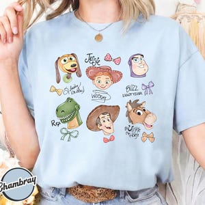 K&ouml;nnte beinhalten: Hellblaues T-Shirt mit Cartoon-Illustrationen von Toy Story-Charakteren. Das Design umfasst Slinky Dog, Jessie, Woody, Buzz Lightyear, Rex und Bullseye das Pferd. Die Namen der Charaktere sind enthalten.