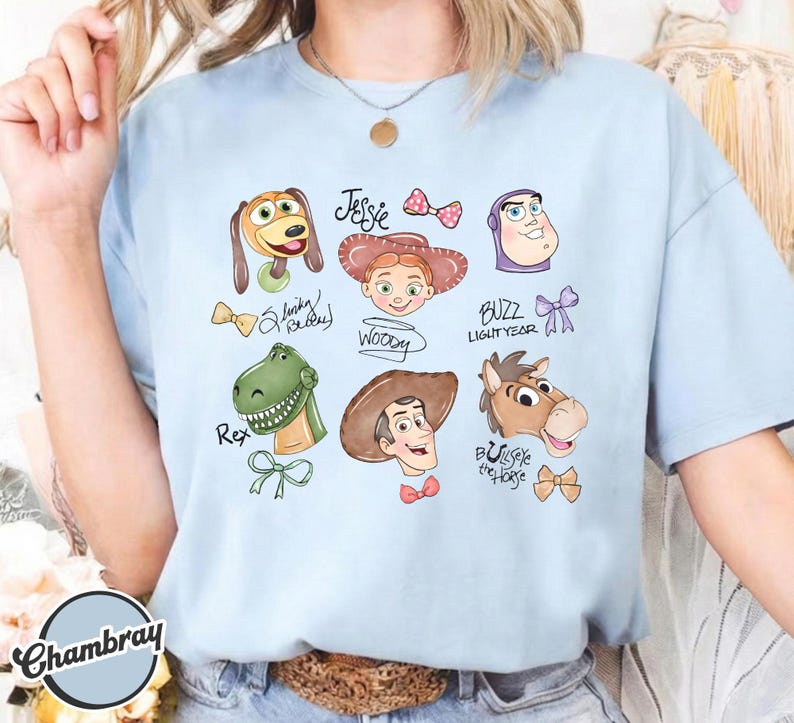 K&ouml;nnte beinhalten: Hellblaues T-Shirt mit Cartoon-Illustrationen von Toy Story-Charakteren. Das Design umfasst Slinky Dog, Jessie, Woody, Buzz Lightyear, Rex und Bullseye das Pferd. Die Namen der Charaktere sind enthalten.