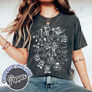 Puede incluir: Camiseta Comfort Colors gris oscuro con un dibujo de contorno blanco de personajes de una popular pel&iacute;cula de animaci&oacute;n. La camiseta tiene cuello redondo y mangas cortas. El logo de Comfort Colors est&aacute; en un c&iacute;rculo en la parte inferior izquierda.