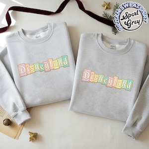 May include: Two light gray sweatshirts with the word "Disneyland" embroidered in pastel colors. The sweatshirts are made of a soft material and have a crew neck. The image also includes a small gold star and a sprig of greenery.