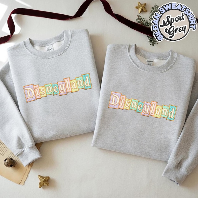 May include: Two light gray sweatshirts with the word "Disneyland" embroidered in pastel colors. The sweatshirts are made of a soft material and have a crew neck. The image also includes a small gold star and a sprig of greenery.