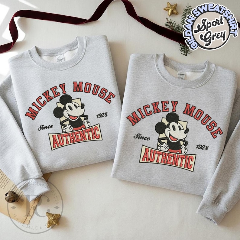 May include: Two light gray sweatshirts featuring a Mickey Mouse graphic. The design includes the words "MICKEY MOUSE" in red arched text, the character's image, and the word "AUTHENTIC" in a red banner. The sweatshirts are folded.