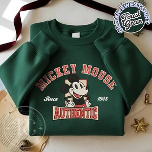 May include: Forest green sweatshirt featuring a Mickey Mouse graphic. The design includes the text "MICKEY MOUSE" in red arched lettering, the word "AUTHENTIC" in a rectangular box, and the year "1928". The sweatshirt has long sleeves.