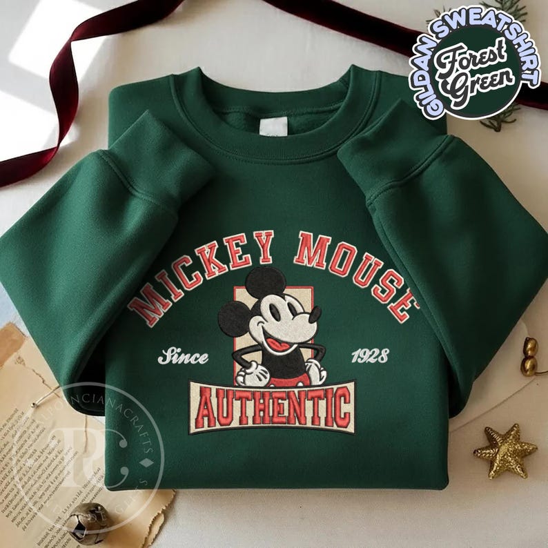 May include: Forest green sweatshirt featuring a Mickey Mouse graphic. The design includes the text "MICKEY MOUSE" in red arched lettering, the word "AUTHENTIC" in a rectangular box, and the year "1928". The sweatshirt has long sleeves.