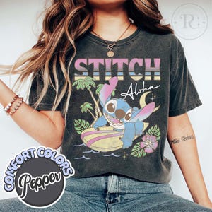 Puede incluir: Camiseta gris oscuro con la palabra "Stitch" encima de un personaje de dibujos animados Stitch surfeando en una tabla de surf. La camiseta tiene la palabra "Aloha" y palmeras en el fondo. La camiseta tambi&eacute;n tiene las palabras "Comfort Colors Pepper" en la parte inferior izquierda.