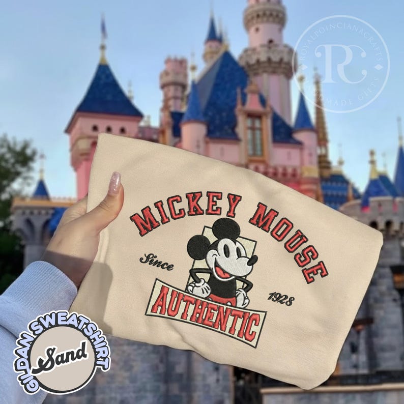 May include: A sand-colored sweatshirt featuring a Mickey Mouse graphic. The design includes the words "MICKEY MOUSE" in red, with "Since 1928" and "AUTHENTIC" also displayed. The sweatshirt is held in front of a castle.