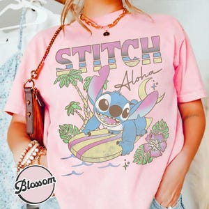 Puede incluir: Camiseta rosa con un gr&aacute;fico colorido de Stitch surfeando, con las palabras "STITCH Aloha". El dise&ntilde;o incluye palmeras, una luna creciente y flores. La camiseta es de color rosa claro.