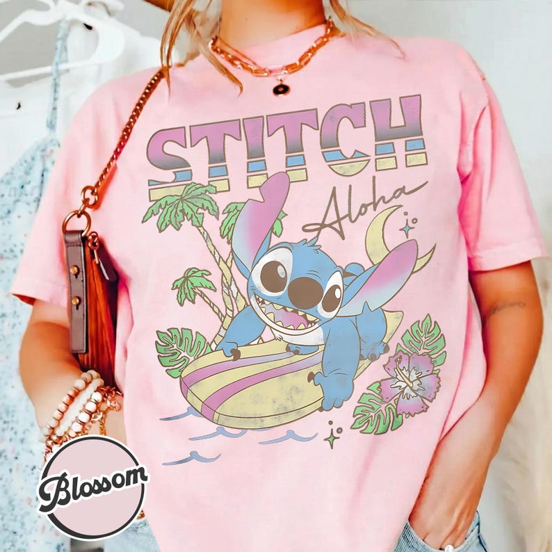 Puede incluir: Camiseta rosa con un gr&aacute;fico colorido de Stitch surfeando, con las palabras "STITCH Aloha". El dise&ntilde;o incluye palmeras, una luna creciente y flores. La camiseta es de color rosa claro.