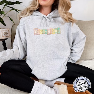 May include: A light gray hooded sweatshirt with the word "Disneyland" embroidered across the chest in pastel colors. The sweatshirt is a Sport Grey Gildan brand.