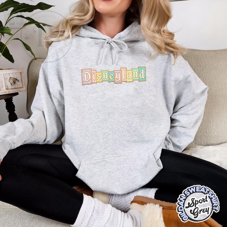 May include: A light gray hooded sweatshirt with the word "Disneyland" embroidered across the chest in pastel colors. The sweatshirt is a Sport Grey Gildan brand.