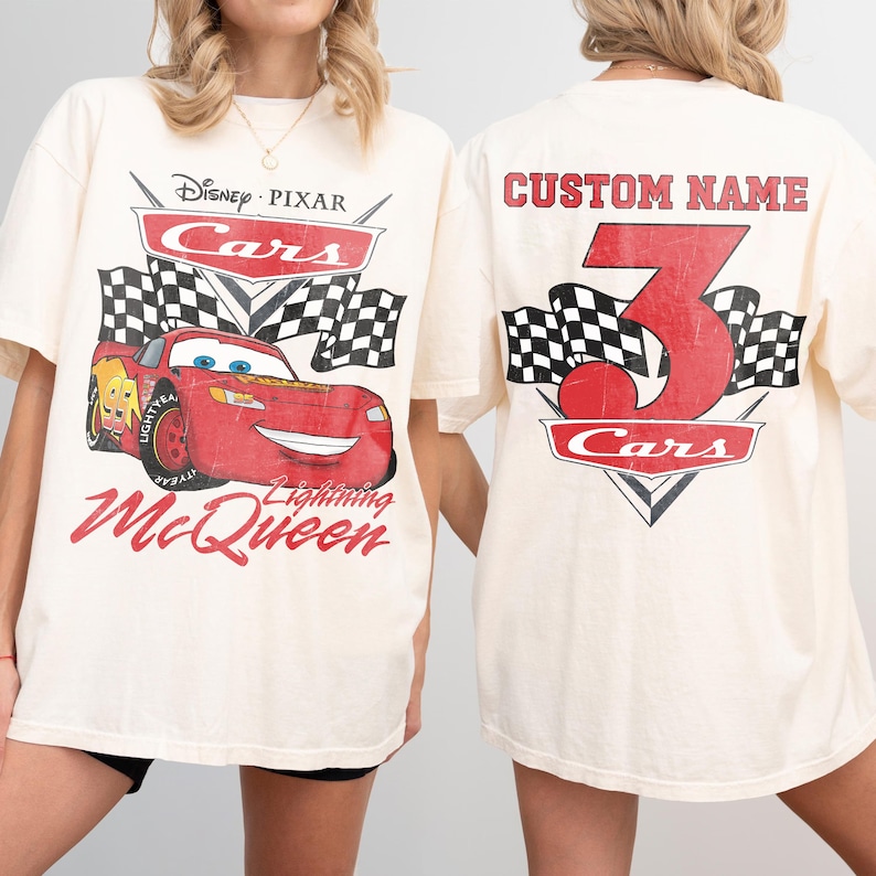 Może przedstawiać: Kremowe koszulki z filmu Auta Disneya Pixara. Prz&oacute;d przedstawia Zygzaka McQueena z szachownicami i napisem "Lightning McQueen". Tył przedstawia dużą czerwoną cyfrę 3, słowo "Cars" i napis "Custom Name."