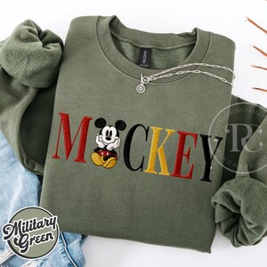 以下が含まれることがあります： 赤、黄、黒で「ミッキー」の文字が刺繍されたミリタリーグリーンのスウェットシャツ。漫画のキャラクターが描かれています。シルバ&mdash;のネックレスが上にかけられています。スウェットシャツはジーンズの上に折りたたまれています。