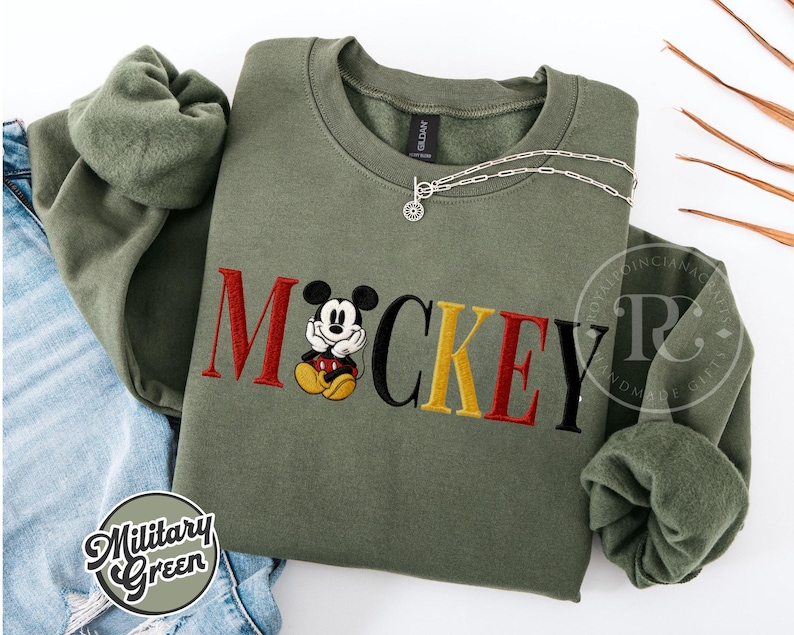 以下が含まれることがあります： 赤、黄、黒で「ミッキー」の文字が刺繍されたミリタリーグリーンのスウェットシャツ。漫画のキャラクターが描かれています。シルバ&mdash;のネックレスが上にかけられています。スウェットシャツはジーンズの上に折りたたまれています。