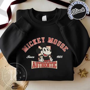 May include: Black sweatshirt featuring a Mickey Mouse graphic. The design includes the words "MICKEY MOUSE" in red arched text, the phrase "Since 1928", and the word "AUTHENTIC" in a rectangular banner. The sweatshirt has a crew neck and long sleeves.