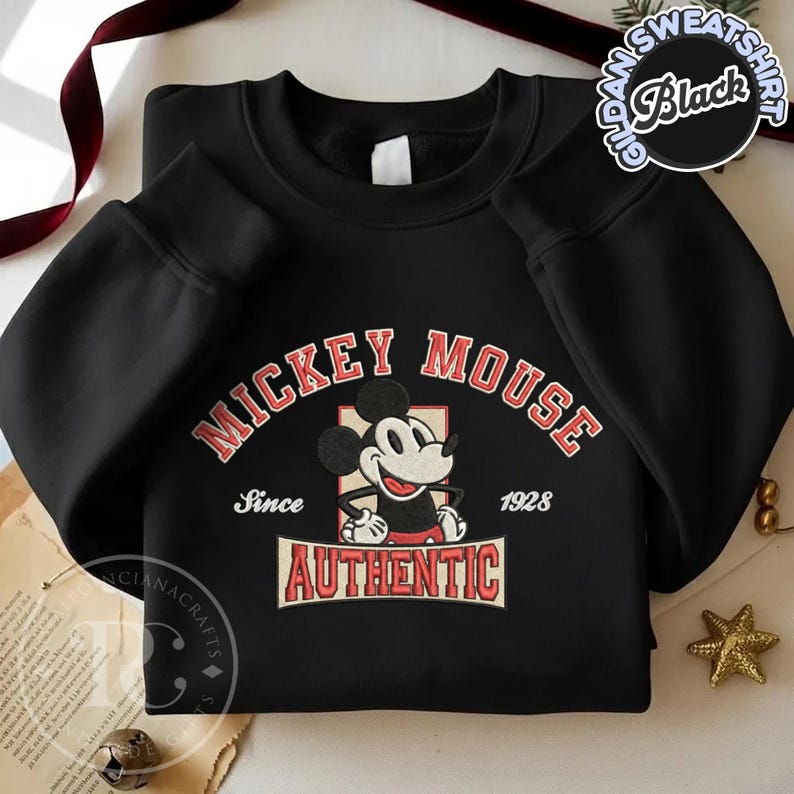 May include: Black sweatshirt featuring a Mickey Mouse graphic. The design includes the words "MICKEY MOUSE" in red arched text, the phrase "Since 1928", and the word "AUTHENTIC" in a rectangular banner. The sweatshirt has a crew neck and long sleeves.
