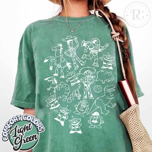 Camiseta unisex de Disney Pixar Toy Story Doodles Comfort Colors, camiseta de dibujos animados de Woody y Buzz Lightyear, camiseta a juego para regalar en un viaje familiar a Disneyland. Light Green