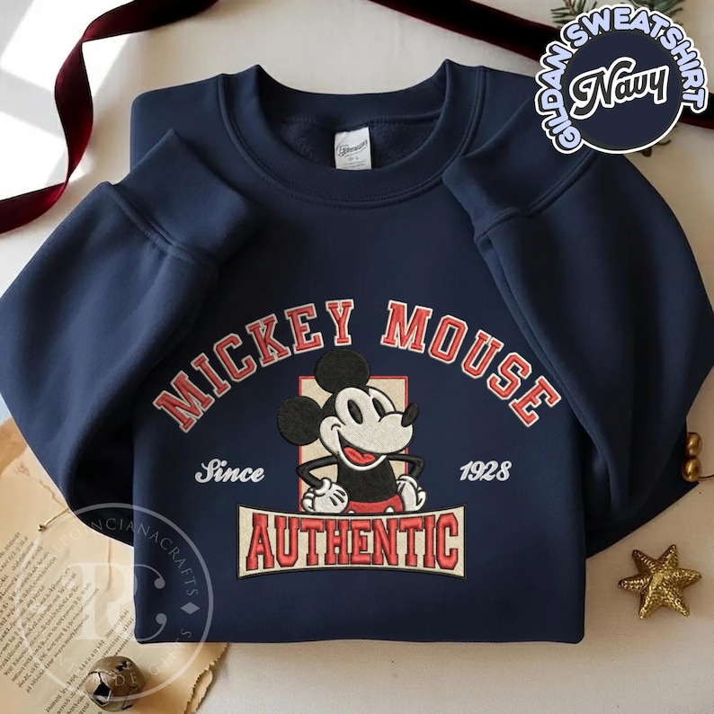 May include: Navy blue sweatshirt featuring a Mickey Mouse graphic. The design includes the words "MICKEY MOUSE" in red, "Since 1928", and "AUTHENTIC". The sweatshirt is a Gildan brand.