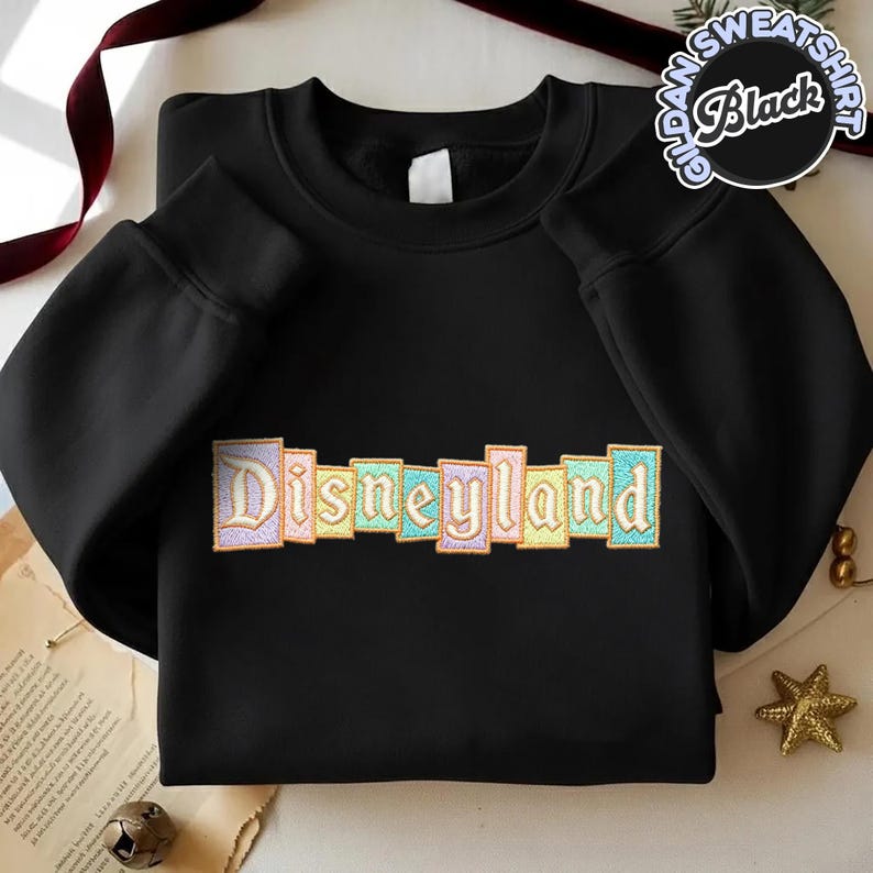 May include: Black crewneck sweatshirt with the word "Disneyland" embroidered across the chest in pastel colors. The sweatshirt has long sleeves and a round neckline. A circular graphic on the upper right reads "Gildan Sweatshirt Black".