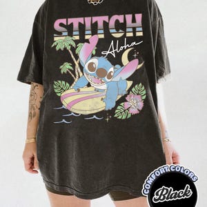 Puede incluir: Camiseta negra con Stitch de Lilo & Stitch surfeando en una tabla de surf. La camiseta tiene las palabras "STITCH Aloha" en una fuente retro colorida. La camiseta es de Comfort Colors.
