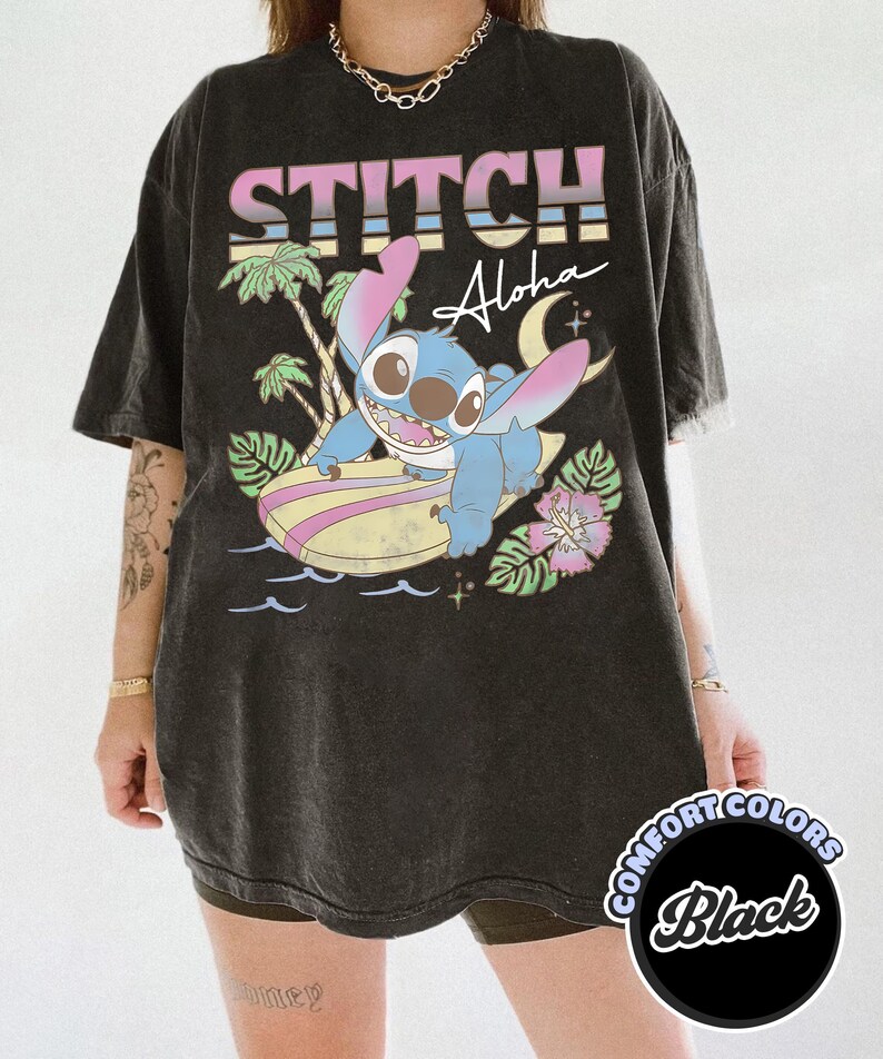 Puede incluir: Camiseta negra con Stitch de Lilo & Stitch surfeando en una tabla de surf. La camiseta tiene las palabras "STITCH Aloha" en una fuente retro colorida. La camiseta es de Comfort Colors.