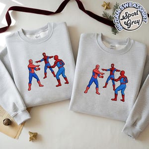 Puede incluir: Dos sudaderas grises claras con un dise&ntilde;o de meme de Spider-Man. El dise&ntilde;o presenta tres figuras de Spider-Man se&ntilde;al&aacute;ndose entre s&iacute;. Las sudaderas tienen mangas largas y cuello redondo. El texto "Gildan Sweatshirt Sport Grey" est&aacute; impreso en la parte superior derecha.