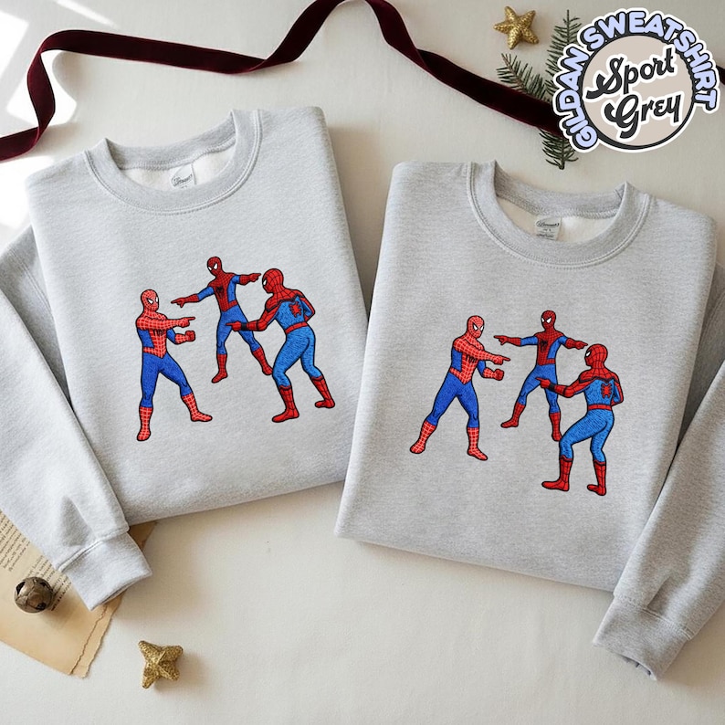 Puede incluir: Dos sudaderas grises claras con un dise&ntilde;o de meme de Spider-Man. El dise&ntilde;o presenta tres figuras de Spider-Man se&ntilde;al&aacute;ndose entre s&iacute;. Las sudaderas tienen mangas largas y cuello redondo. El texto "Gildan Sweatshirt Sport Grey" est&aacute; impreso en la parte superior derecha.