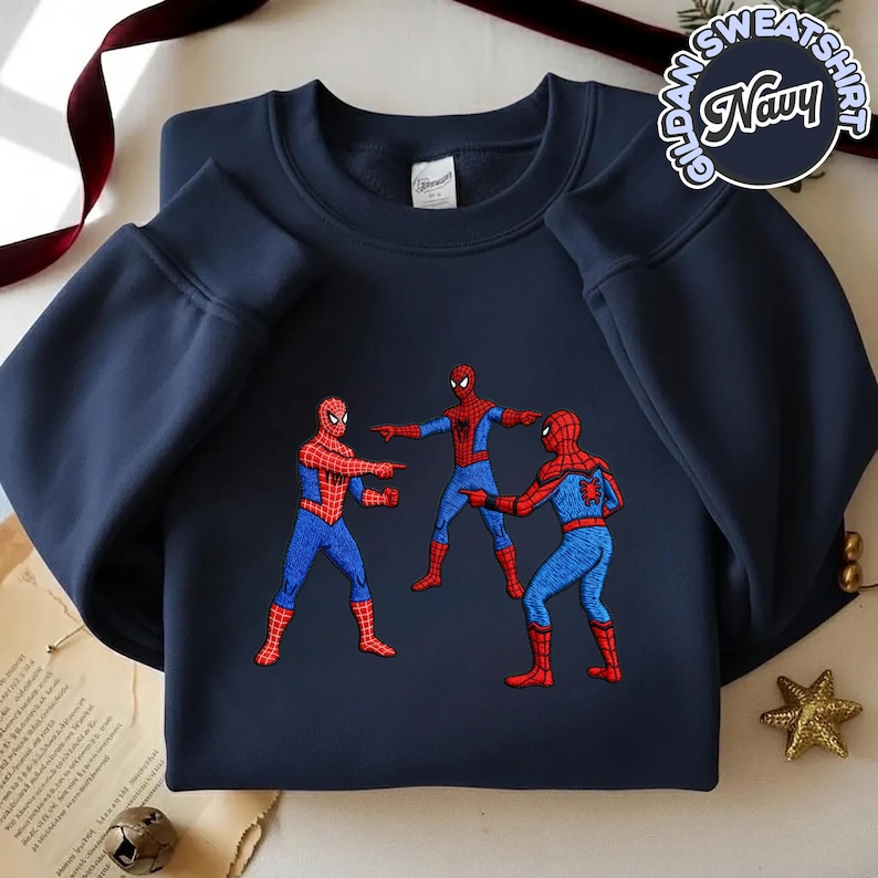 Puede incluir: Sudadera azul marino con un dise&ntilde;o bordado de tres personajes de Spider-Man se&ntilde;al&aacute;ndose. La sudadera tiene mangas largas y cuello redondo. El texto "Gildan Sweatshirt Navy" est&aacute; impreso en la esquina superior derecha.