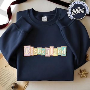 May include: Navy blue Gildan sweatshirt with the word "Disneyland" embroidered in pastel colors. The sweatshirt has a crew neck and long sleeves. A navy blue circle with the words "Gildan Sweatshirt Navy" is in the upper right corner.