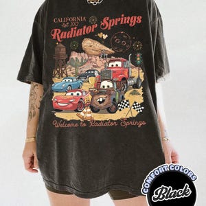 Pode incluir: Camiseta preta com um gr&aacute;fico dos personagens do filme Carros e o texto "California Est. 2012 Radiator Springs Welcome to Radiator Springs." A camisa &eacute; da marca Comfort Colors.