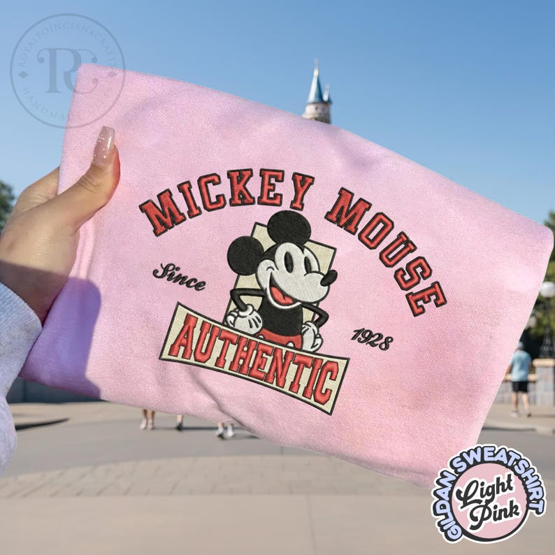 May include: Light pink sweatshirt featuring a Mickey Mouse graphic. The design includes the words "Mickey Mouse" in red, the word "Authentic" in a banner, and the year "1928". The sweatshirt is a light pink color.