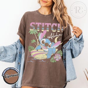 Puede incluir: Camiseta color espresso con un gr&aacute;fico de Stitch surfeando, con la palabra "STITCH" arriba y "Aloha" abajo. El dise&ntilde;o incluye palmeras, olas y una tabla de surf. La camiseta es de Comfort Colors.