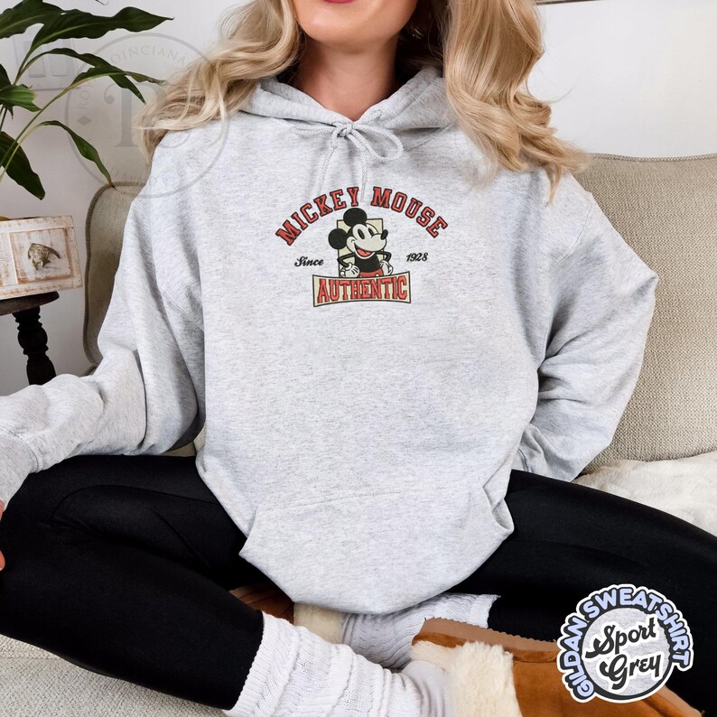 May include: A light gray hoodie featuring a Mickey Mouse graphic. The graphic includes the text "Mickey Mouse Since 1928 Authentic" in red and black. The hoodie is made of a soft material and has a drawstring hood.