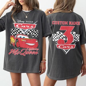 Może przedstawiać: Ciemnoszara koszulka z motywem Disney Pixar Cars z Zygzakiem McQueen. Grafika z przodu zawiera logo Cars i flagi w szachownicę. Z tyłu znajduje się numer 3 oraz napis "Custom Name".