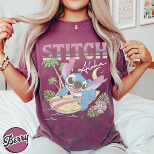 Puede incluir: Camiseta granate con un dise&ntilde;o de Stitch de Lilo & Stitch surfeando, con la frase "STITCH Aloha" en una fuente retro. El dise&ntilde;o incluye palmeras, una luna creciente y flores tropicales.