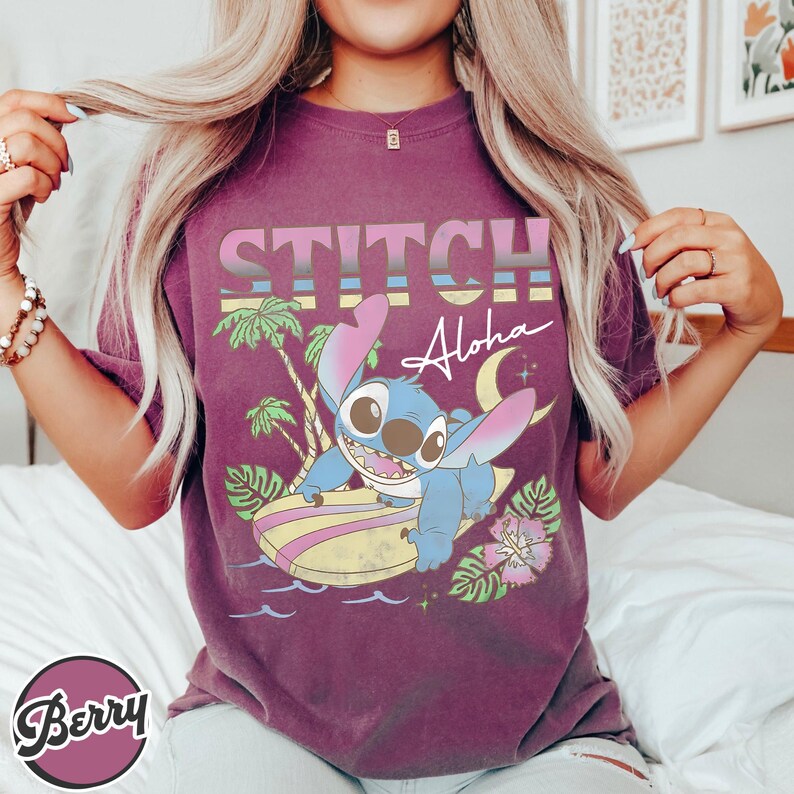 Puede incluir: Camiseta granate con un dise&ntilde;o de Stitch de Lilo & Stitch surfeando, con la frase "STITCH Aloha" en una fuente retro. El dise&ntilde;o incluye palmeras, una luna creciente y flores tropicales.