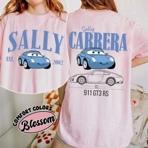 May include: Pink t-shirt featuring a light blue cartoon car with the text "SALLY CARRERA" and "EST. 2002". The back of the shirt also has a car graphic and the text "911 GT3 RS". The shirt has the "Comfort Colors Blossom" logo.