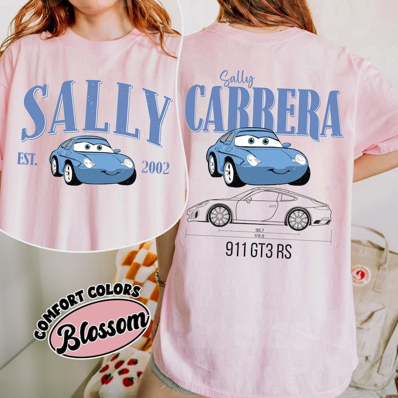 May include: Pink t-shirt featuring a light blue cartoon car with the text "SALLY CARRERA" and "EST. 2002". The back of the shirt also has a car graphic and the text "911 GT3 RS". The shirt has the "Comfort Colors Blossom" logo.