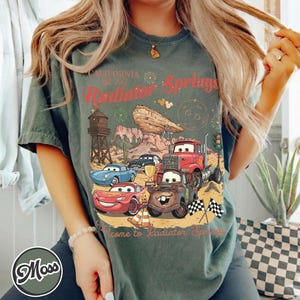 Pode incluir: Camiseta verde com um gr&aacute;fico de Radiator Springs, com carros de desenho animado, uma torre de &aacute;gua e o texto "California Radiator Springs". A camisa tem um visual vintage e desgastado.