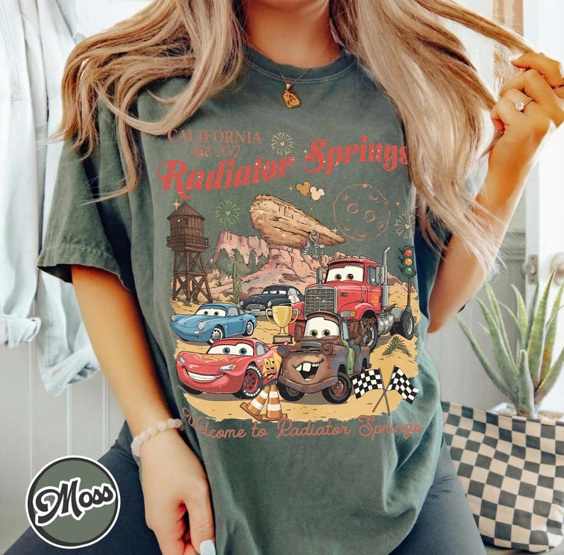Pode incluir: Camiseta verde com um gr&aacute;fico de Radiator Springs, com carros de desenho animado, uma torre de &aacute;gua e o texto "California Radiator Springs". A camisa tem um visual vintage e desgastado.