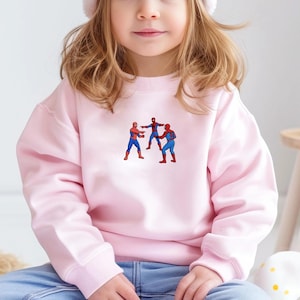Puede incluir: Sudadera rosa claro con un gr&aacute;fico de tres figuras de Spider-Man con trajes rojos y azules. Las figuras est&aacute;n en poses de acci&oacute;n. La sudadera est&aacute; hecha de un material suave.