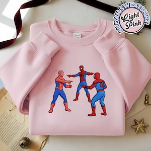 Puede incluir: Sudadera Gildan rosa claro con un dise&ntilde;o bordado de tres figuras de Spider-Man se&ntilde;al&aacute;ndose. La sudadera tiene mangas largas y cuello redondo. El texto "Light Pink" est&aacute; en un dise&ntilde;o circular en la parte superior derecha.