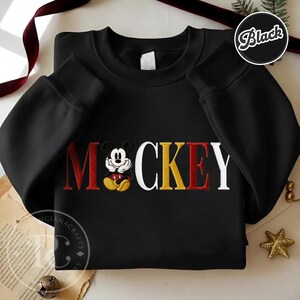 以下が含まれることがあります： 赤、黄、白で「ミッキー」の文字が刺繍された黒のクルーネックのスウェットシャツ。ミッキーマウスのグラフィックが特徴です。スウェットシャツは長袖で、丸いネックラインです。