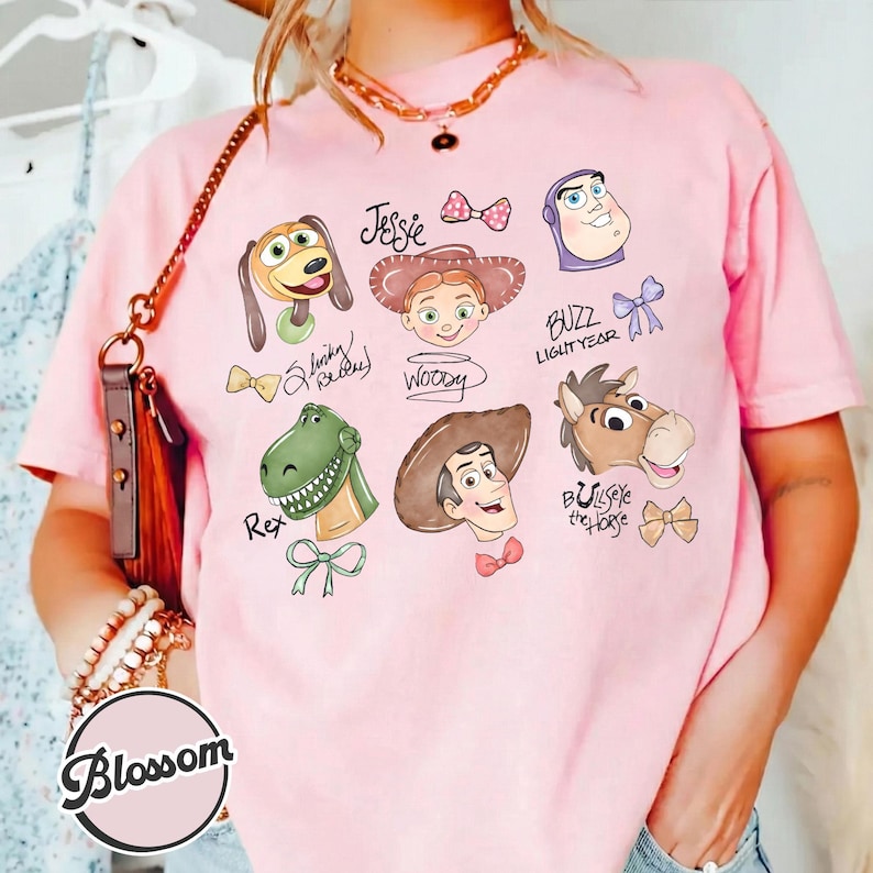 K&ouml;nnte beinhalten: Hellrosa T-Shirt mit Cartoon-Illustrationen von Toy Story-Charakteren, darunter Woody, Buzz Lightyear und Jessie. Das Wort "Blossom" ist unten links auf dem Shirt aufgedruckt.