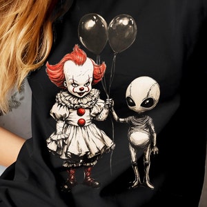 May include: Black t-shirt featuring a cartoon illustration of a clown and an alien holding black balloons. The clown has red hair and a white ruffled dress. The alien is gray with a large head and thin body.
