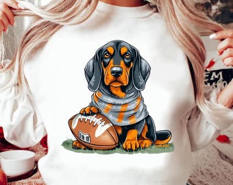 Puede incluir: Sudadera blanca con una imagen de dibujos animados de un dachshund negro y marrón sosteniendo un balón de fútbol marrón y blanco. El dachshund lleva una bufanda gris y naranja.
