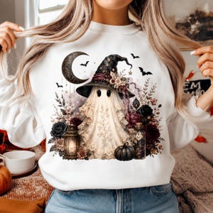 May include: A white crewneck sweatshirt featuring a graphic of a ghost wearing a witch hat, surrounded by dark flowers, a lantern, a pumpkin, and a crescent moon. The ghost is draped in lace-like fabric, with small bats flying in the background.