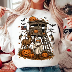 May include: A white sweatshirt featuring a graphic print of a wooden hunting stand, a deer, ducks, and a ghost wearing a hat that says CANDY HUNTER and holding a rifle. The design includes pumpkins, bats, autumn leaves, and an American flag.